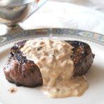 On a white plate is served a grilled steak with a creamy peppercorn sauce on top.