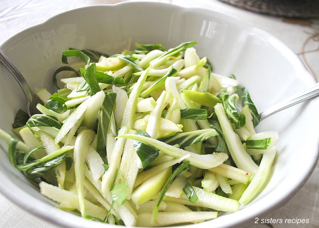 A white salad bowl with sliced apples, and sliced Bok Choy. 