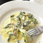A white dinner plate with a serving of ravioli topped with a cream and asparagus sauce.