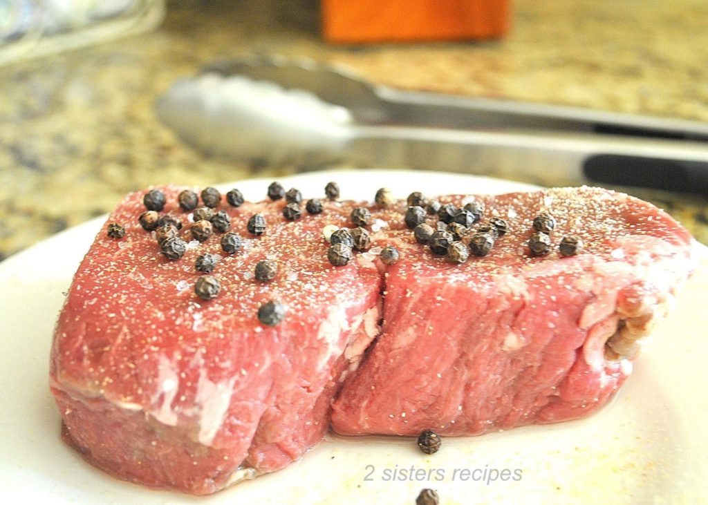 A raw steak on a plate with peppercorns on top. by 2sistersrecipes.com
