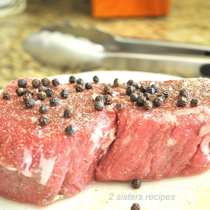 A raw steak on a plate with peppercorns on top. by 2sistersrecipes.com