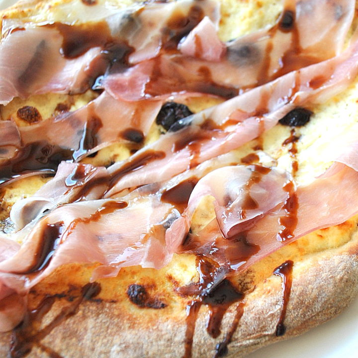 A baked pizza topped with prosciutto and cherries and balsamic glaze drizzled across the top.