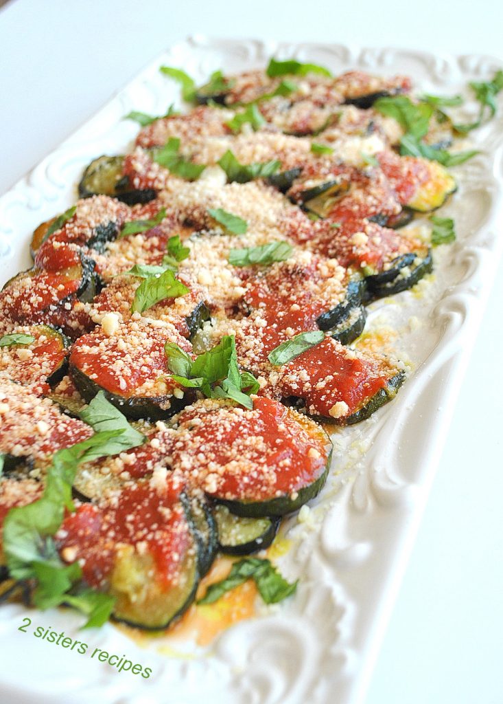 A long white platter filled with zucchini disks roasted and topped with tomato sauce and grated cheese. 