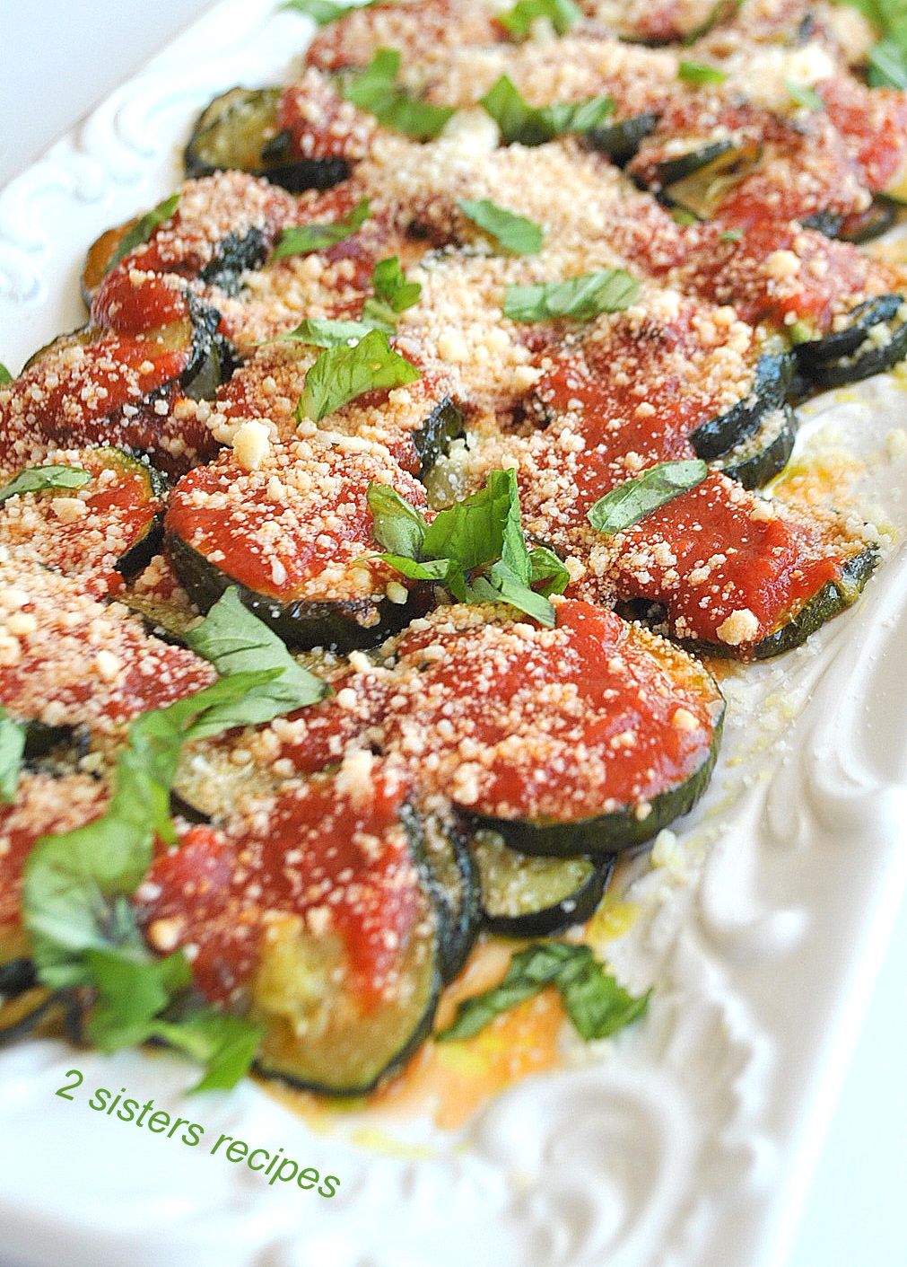 A long white serving platter with sliced zucchini disks topped with tomato sauce, grated cheese and fresh basil.