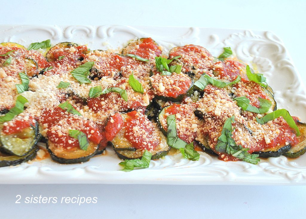 A white platter with a serving of zucchini disks, topped with tomato sauce, and grated cheese, and ribbons of basil. 