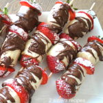 A white platter with skewers filled with sliced bananas and strawberries with chocolate drizzled over them.