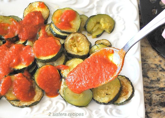 Spooning tomato sauce over the roasted zucchini disks on a white platter.