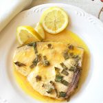 A baked swordfish with lemon sauce served on a white plate, with lemon slices on the side.