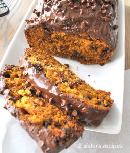 A pumpkin bread sliced on a white platter is covered with chocolate garnash and mini chocolate chips.