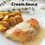 A white plate with a serving of a grilled chicken breast with cream sauce and a side of roasted potatoes.