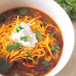 A white bowl of beans and vegetable chili with shredded cheddar, and some sour cream, and cilantro on top.