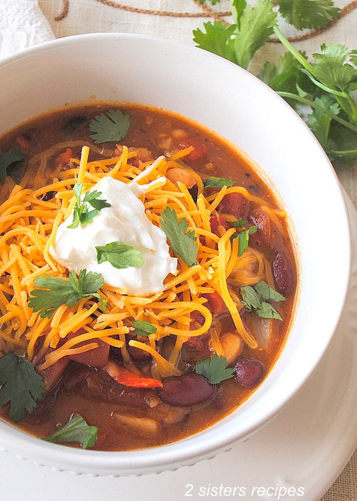 A white bowl of beans and vegetable chili with shredded cheddar, and some sour cream, and cilantro on top.