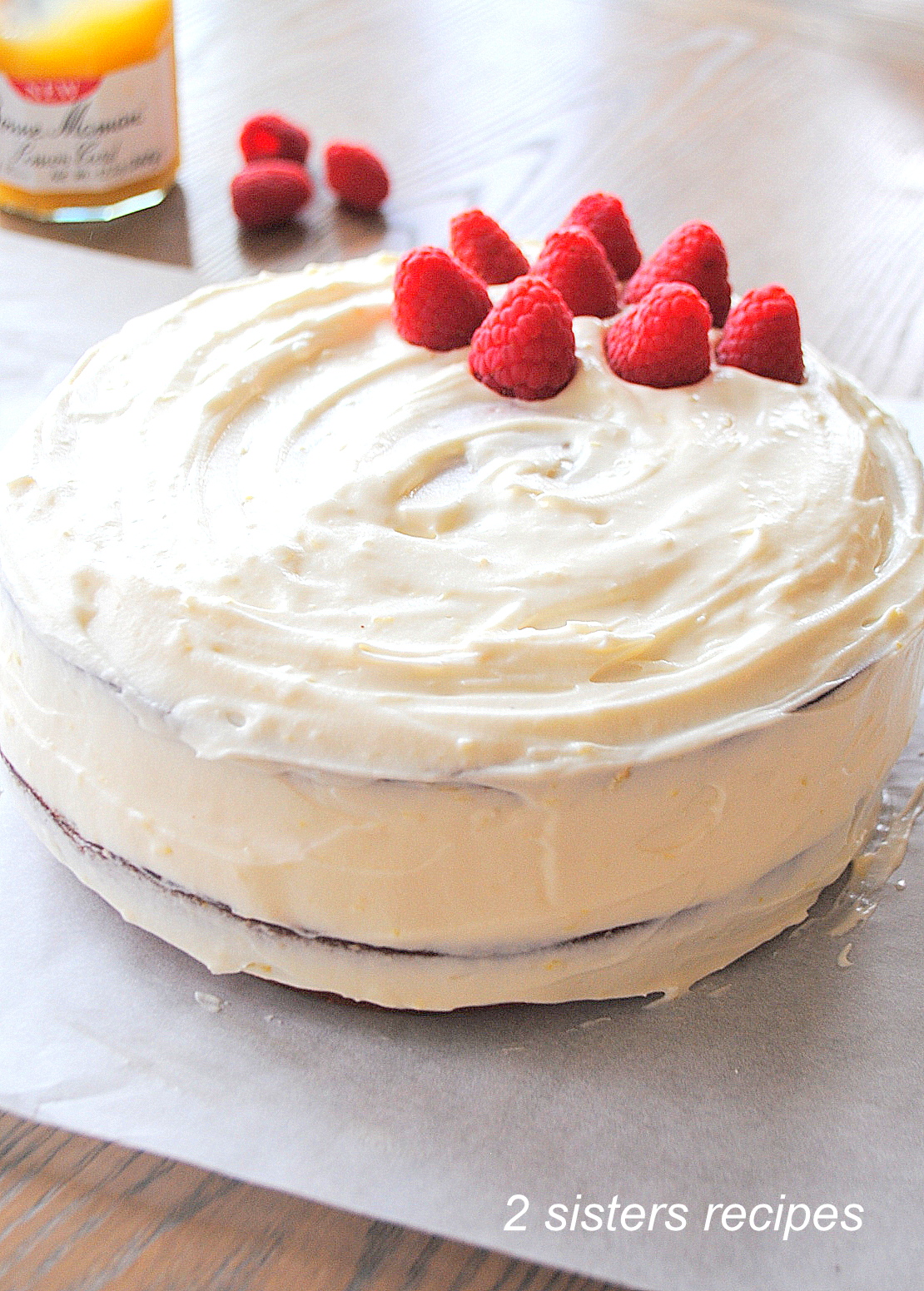 The whole cake with white frosting and fresh raspberries on top. by 2sistersrecipes.com