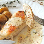 A forkful of chicken with Dijon cream sauce served on a white plate with a side of potatoes.