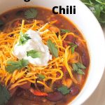 A white bowl filled with a chili, topped with shredded cheddar cheese and dollop of sour cream on top.