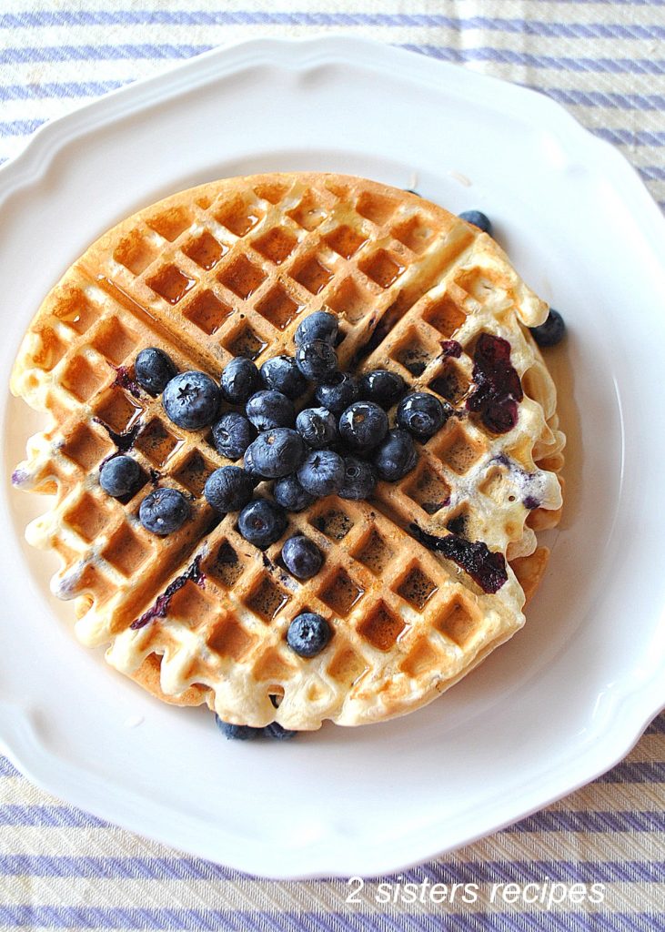 A waffle with fresh blueberries on top on a white plate.