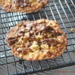 A baked thin baked cookie with a drizzle of melted chocolate on top, set on a black wire rack to cool.