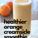 Healthier Orange Creamsicle Smoothis is served in a tall glass with a straw and 2 navel oranges on the side.