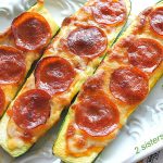 There are 3 zucchini boats topped with sliced pepperoni served on a white platter.