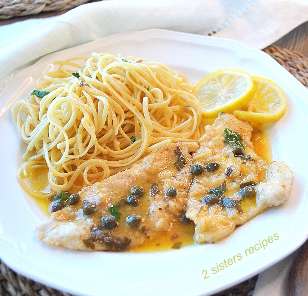 A white dinner plate with a cooked chicken with lemon sauce and capers and a side of spaghetti. 