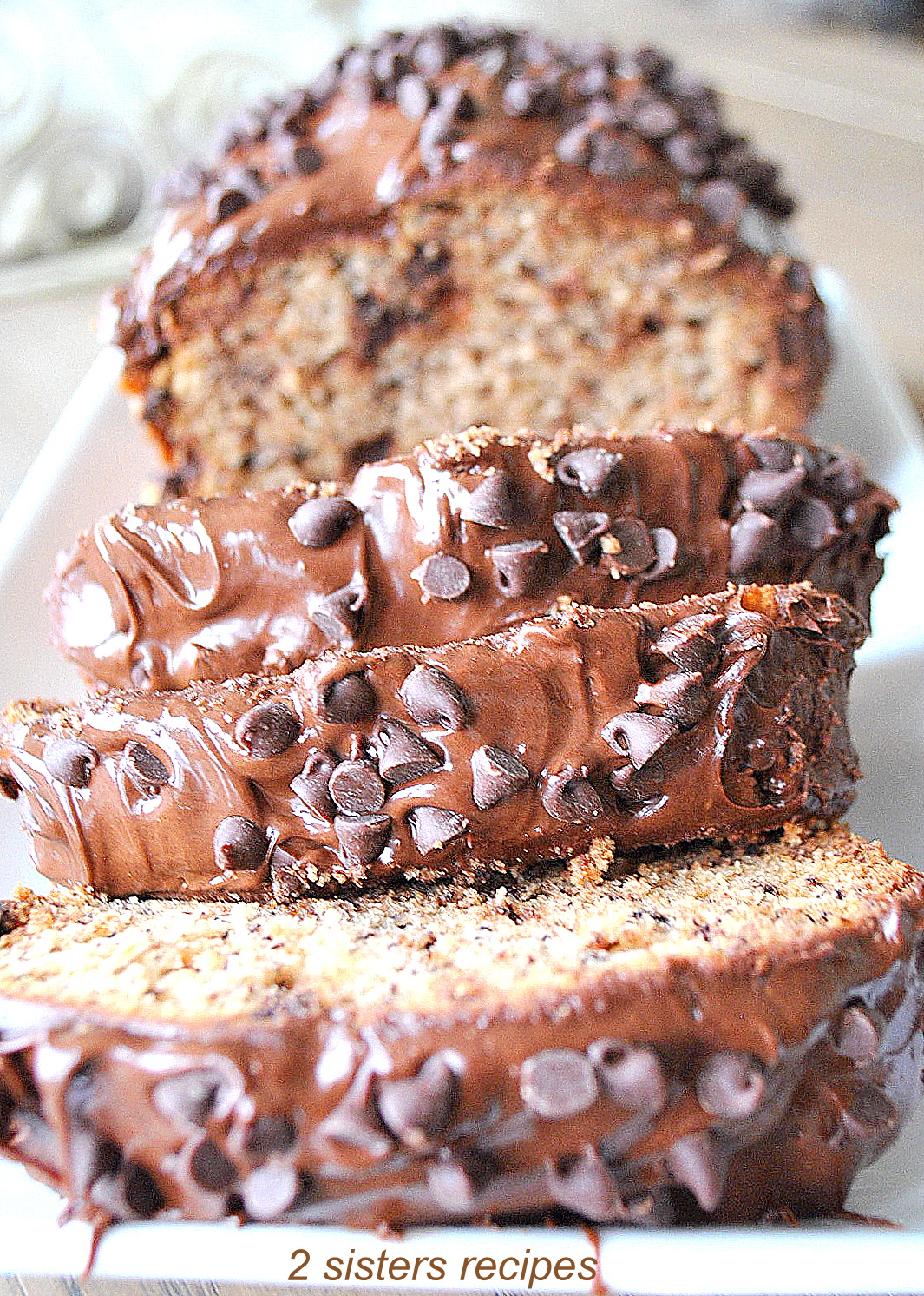 A loaf of bread with chocolate frosting, and mini chocolate chips on top, sliced on a white platter.