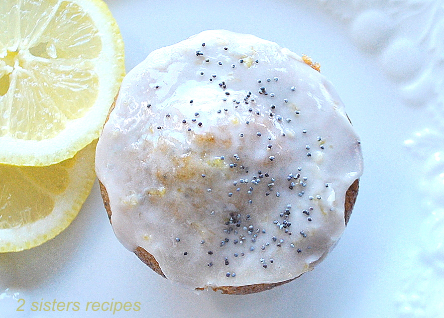 Looking at the top of a glazed muffin with poppy seeds scattered on top, with lemon slices on the side.