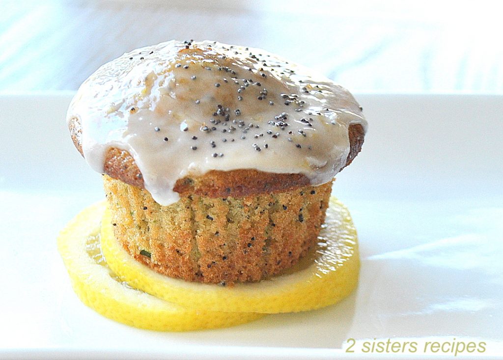 One lemon muffin  topped with icing and poppy seeds, placed on 2 lemon sliceds on a white plate.