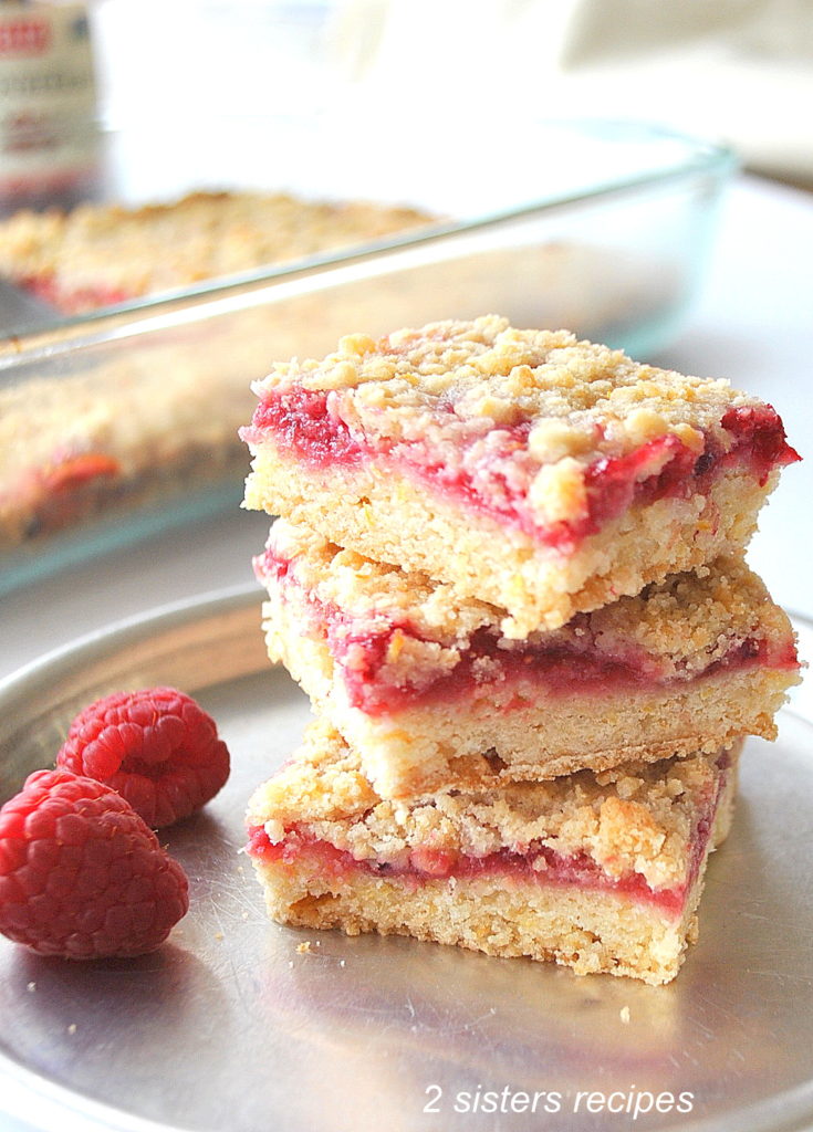 3 Raspberry Lemon Crumble Bars are piled on top of each other served on a silver plat with fresh raspberries on the side.