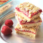 3 raspberry filled bars piled on top of one another with 2 fresh raspberries on the side served on a silver plate.
