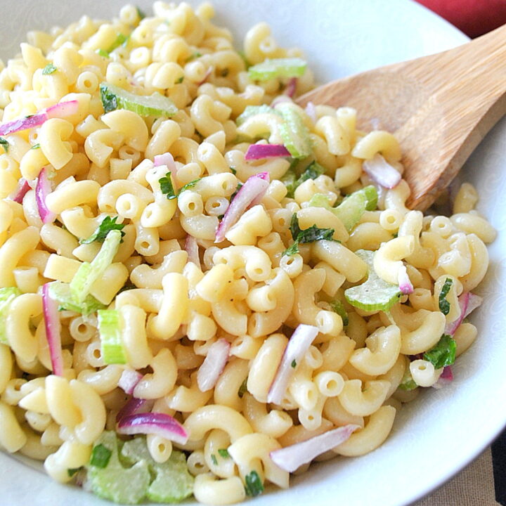 A white bowl filled with elbow pasta with celery and red onion with a wooden spoon in it.