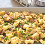 A white casserole dish filled with tortellini, peas, and bacon bits.
