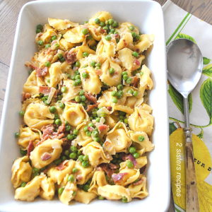 A white rectangular casserole dish filled with tortellini, bits of bacon, and peas.