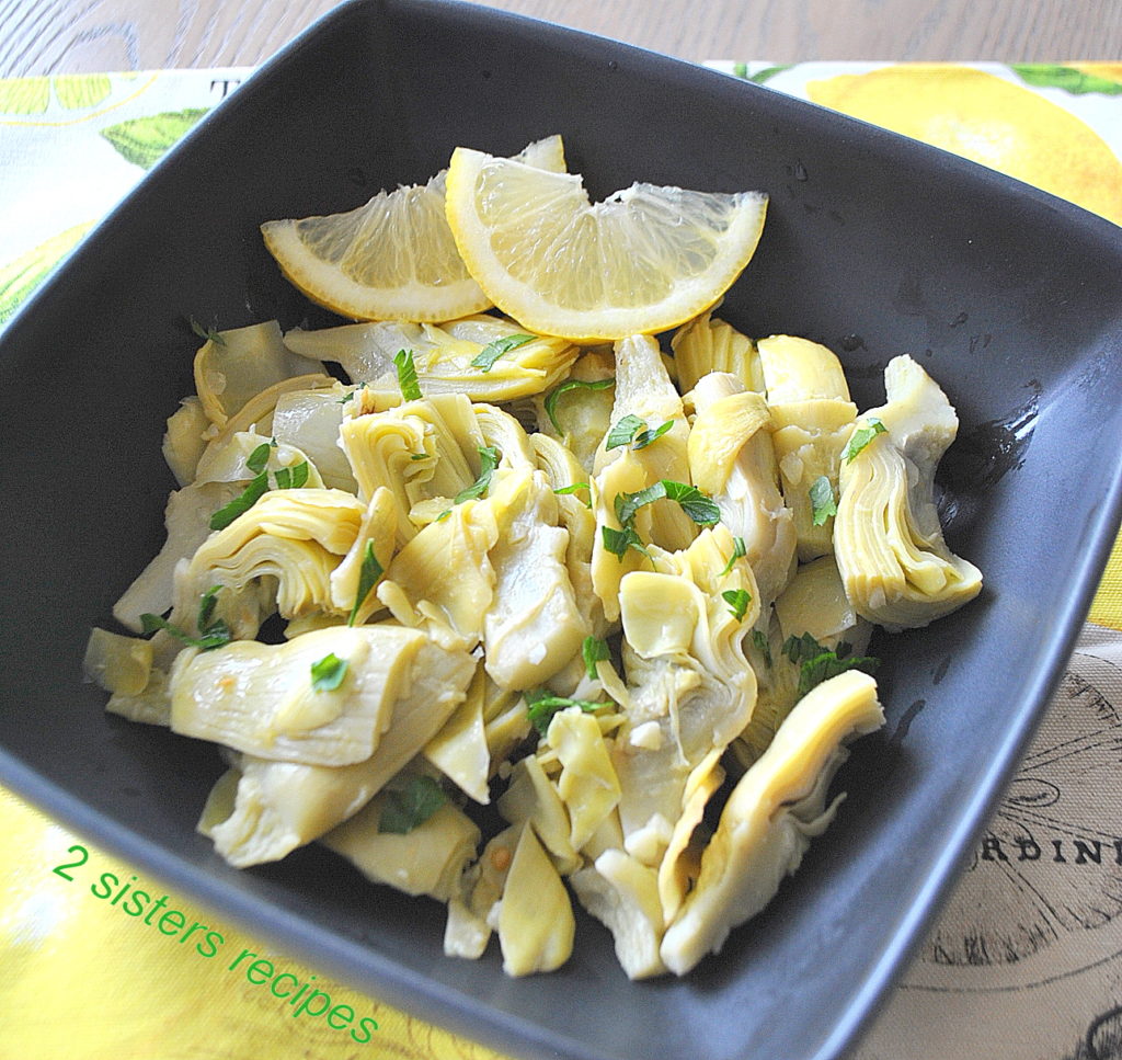 A black square bowl filled with artichoke hearts, chopped parsley and lemon slices on the side.