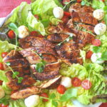 A sliced grilled chicken served on a bed of lettuce with cherry tomatoes and small mozzarella balls.