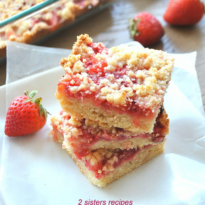A pile of strawberry and pineapple crumble bars served on a white plate, with a fresh strawberry on the side.