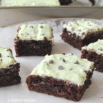 Squares of chocolate brownies topped with a light green icing and mini chocolate chips served on a white platter.