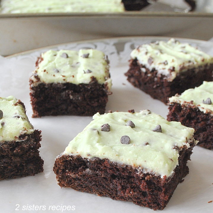 Squares of chocolate brownies topped with a light green icing and mini chocolate chips served on a white platter.