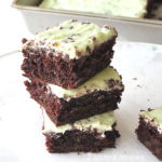Chocolate Chip Zucchini Brownies are piled on top of each other with a light green frosting and mini chocolate chips on top.