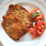 A fried veal cutlet is served on a white plate with a side of a tomato salad.