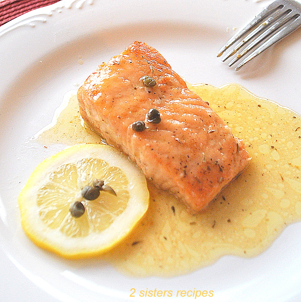 A white dinner plate with a cooked salmon topped with a lemon sauce and capers.
