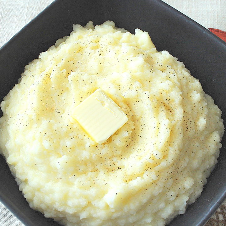 A black square serving bowl filled with mashed potatoes with a slice of butter on top.