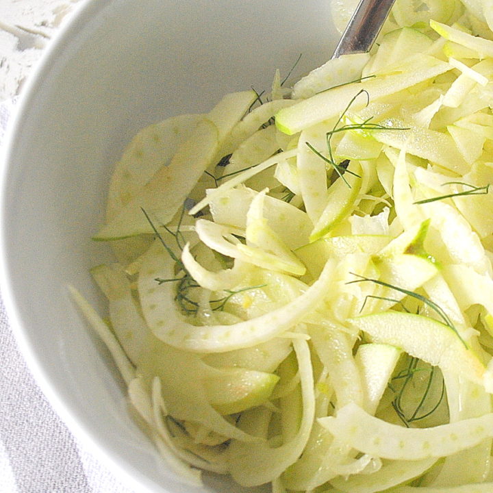 A white salad bowl with a serving spoon inside, along with thinly shavings of apples and fennel.