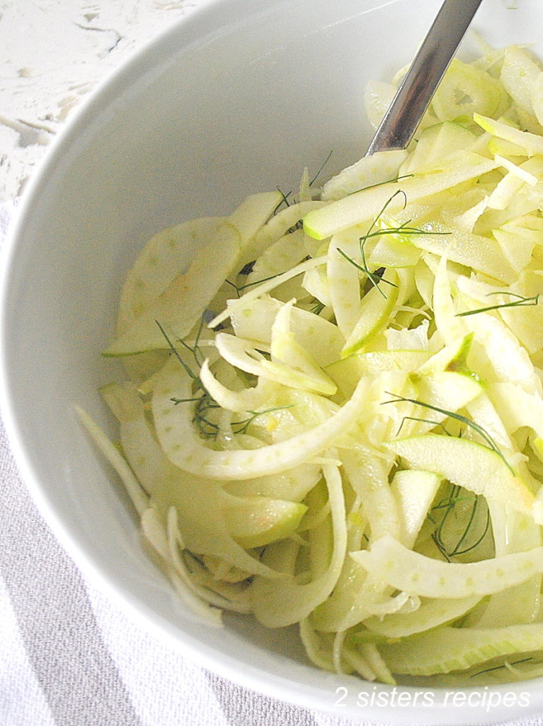 A white salad bowl with a serving spoon inside, along with thinly shavings of apples and fennel.