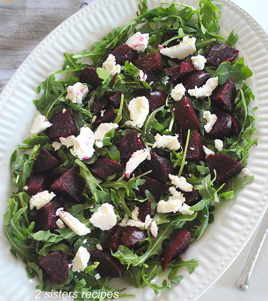 A large white platter filled with scattered arugula greens, chopped beets and chunks of cheese. 