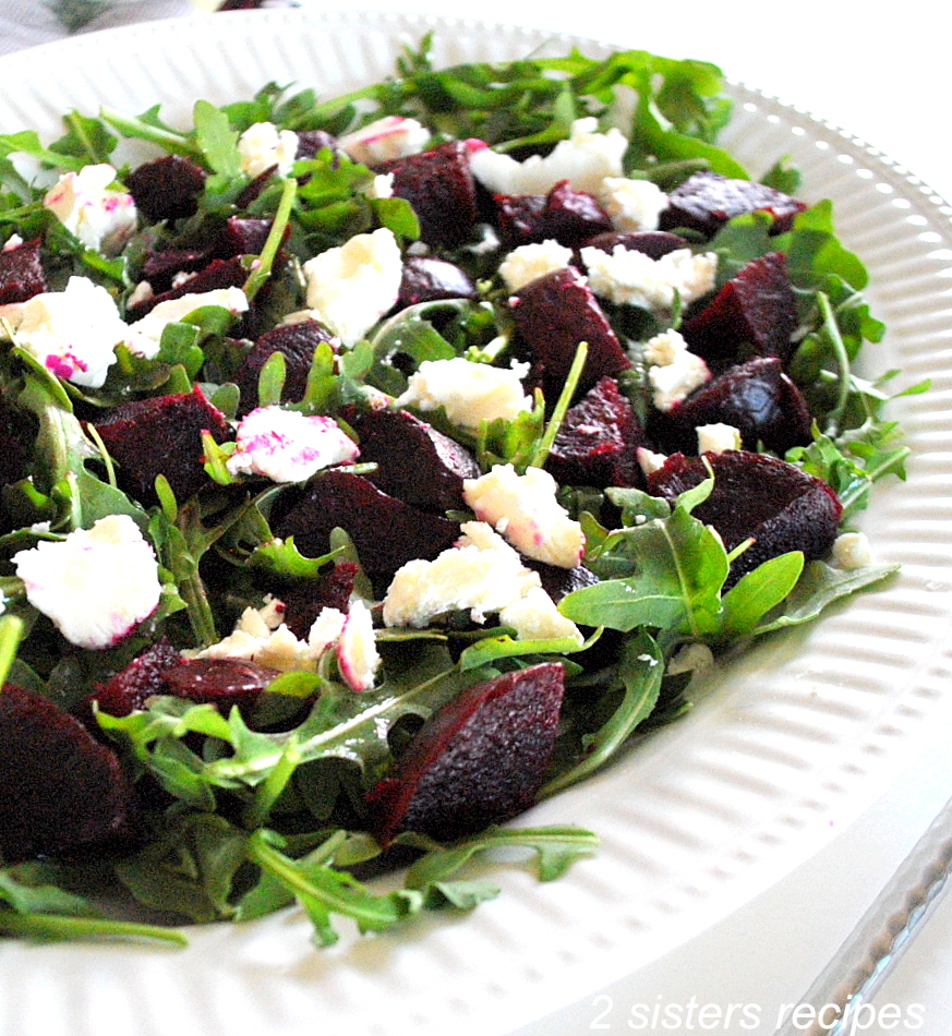 A large white salad platter filled with arugula, chopped beets and chunks of goat cheese on top.