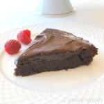 A slice of chocolate cake with ganache on top served on a white plate with 2 raspberries on the side.