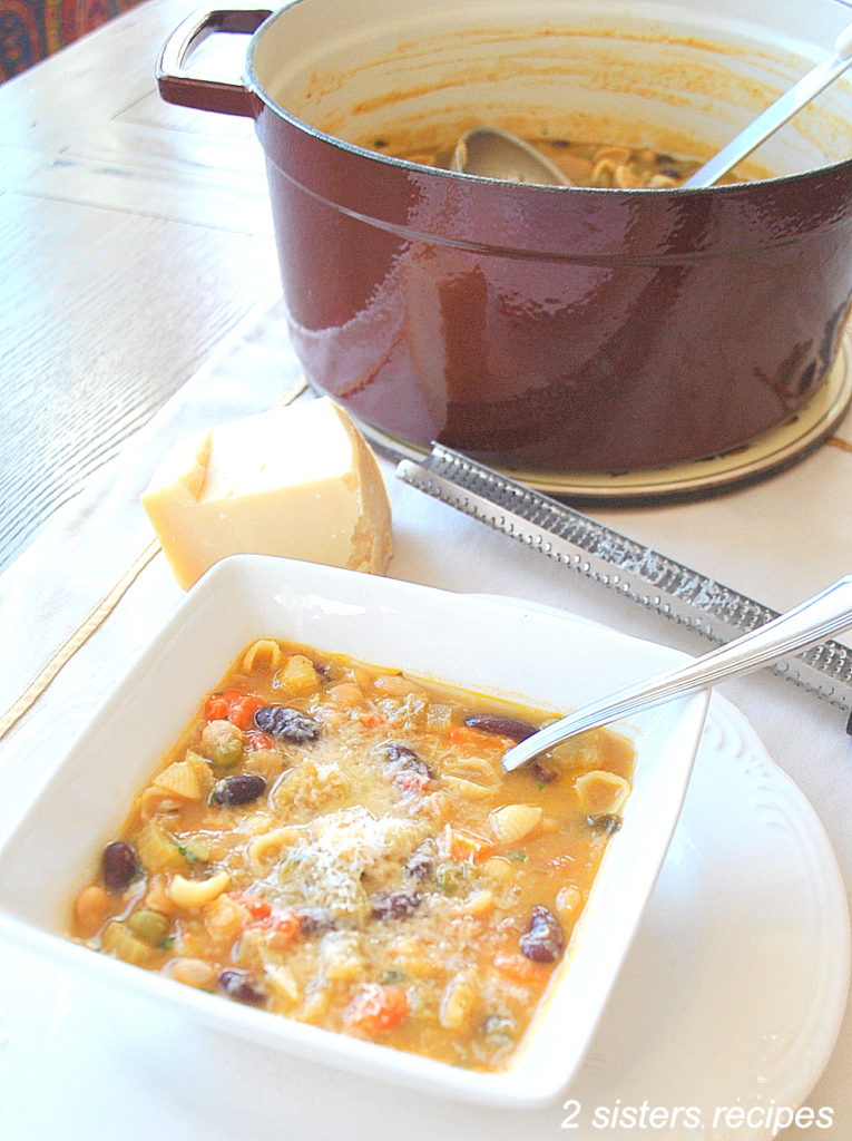 A bowl of vegetable soup on the table with a red Dutch oven pot next to it, and a chunk of parmigiana cheese next to it. 