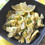 A black square bowl filled with artichoke hearts and lemon slices.