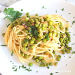 A white plate filled with spaghetti, topped with peas and pancetta.
