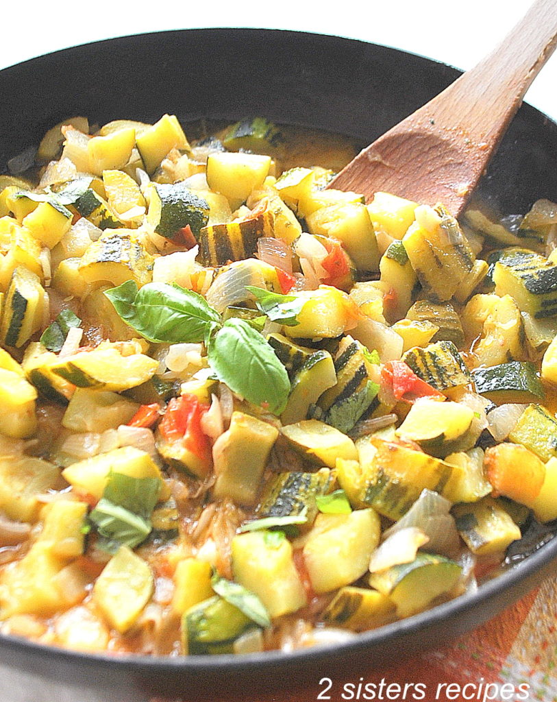 A large skillet loaded with zucchini stew and a wooden spoon inside.